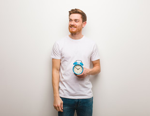 Young redhead man smiling confident and crossing arms, looking up. He is holding an alarm clock.