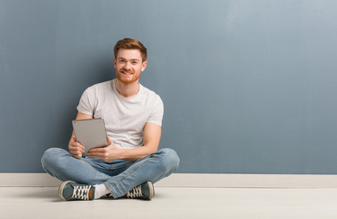 Young redhead student man sitting on the floor cheerful with a big smile. He is holding a tablet. - obrazy, fototapety, plakaty
