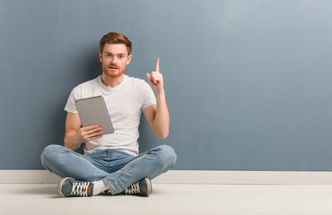 Young redhead student man sitting on the floor showing number one. He is holding a tablet.