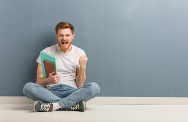 Young redhead student man sitting on the floor surprised and shocked. He is holding books.