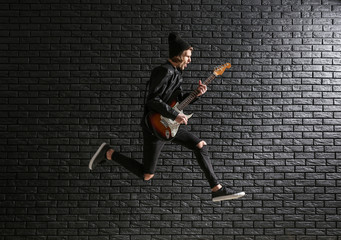 Jumping young man with guitar against dark brick wall