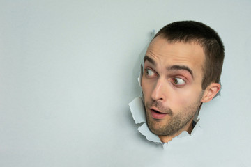 Dazed man making hole in paper, looks aside, opening his mouth, blue background, copy space