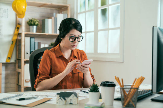 asian woman engineer employee sitting at desk in office checking on blueprint take photo sending message by smart phone. friendly small start up business woman architect working diagrams and graphs - Powered by Adobe