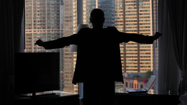 Man in casual jacket stretch oneself, expand hands and breathe, move shoulders and put arms on hips, black silhouette against room window. Mansion house facade seen blurred outdoors