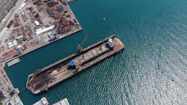 Cargo ship stay in dry floating dock, aerial shot. Large vessel under repair or maintenance. Various steel parts of watercraft stacked on dockyard ground. Camera soar over dock area