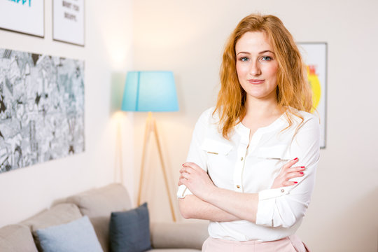 young Caucasian girl standing in the office and folded her arms across her chest. Charming young girl works inside in bright office, coworking room. The girl has blond red hair and white shirt