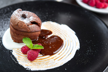Chocolate pudding lava cake with vanilla cream, raspberry and mint on a black plate. Sweet muffins for dessert