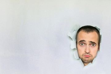 Sad guy making hole in paper, looking at the camera, grieving, pursing his lips, blue background with a copy of the space