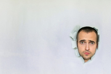 Handsome young guy making hole in paper, looking at the camera, grieving, blue background, copy space