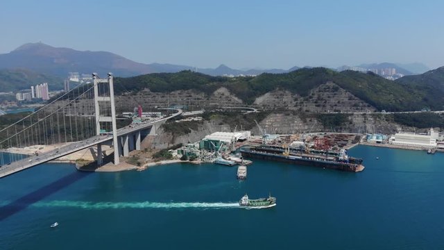 Large Tsing Ma suspension bridge landing at Tsing Yi island, dock yards on shore, dry dock vessel on water, aerial shot, camera fly towards land side. Ship sail along Ma Wan channel, green top of hill