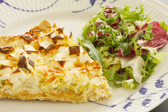 A slice of quiche with leek and feta or goats cheese on a plate with crispy leaf green salad. Vegetarian food in close up image.