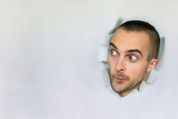 Curious man making hole in paper, interested looks aside, surprise, blue background, copy space