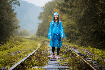 Girl traveller wearing blue jacket and go by forest railway. Autumn and raining season with dark green tones while girl in blue rain jacket walks in forest, fog