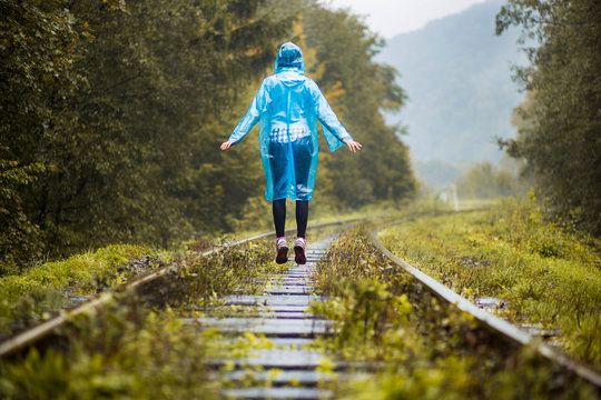 Girl Traveller Wearing Blue Jacket And Go By Forest Railway. Autumn And Raining Season With Dark Green Tones While Girl In Blue Rain Jacket Walks In Forest, Fog