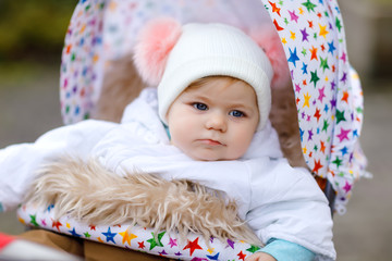 Cute little beautiful baby girl sitting in the pram or stroller on autumn day. Happy healthy child going for a walk on fresh air in warm clothes. Baby with in colorful clothes and hat with bobbles