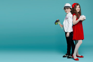 Portrait of two children standing with back to back. Handsome boy in elegant suit with eye glasses, hat and beautiful brunette girl in red dress posing in studio, isolated on turquoise background.
