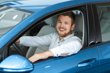 Driving is his passion. Happy handsome man smiling cheerfully to the camera while sitting in his new car at the dealership