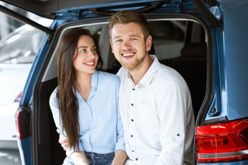 Happy about their buy. Beautiful young loving woman smiling at her handsome happy husband as he is laughing to the camera while sitting together in a trunk of a new car