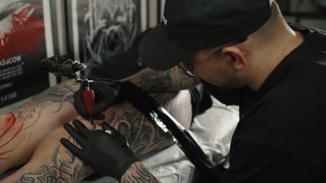 Master man tattoo in glasses draws the color paint on the clients tattoo. Tattoo artist holding tattoo machine in black sterile gloves