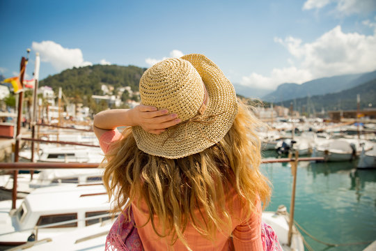 Portrait of woman with light curly hair, rear view,  holding straw hat with hand. Sunny harbor, boats and yachts, green mountains on background. Enjoying life, happy traveling,