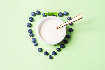 Bowl of natural yogurt and fresh blueberries in shape of heart on pastel green background. Healthy breakfast concept