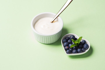 Bowl of natural yogurt and fresh blueberries in heart shaped plate on pastel green background. Healthy breakfast concept