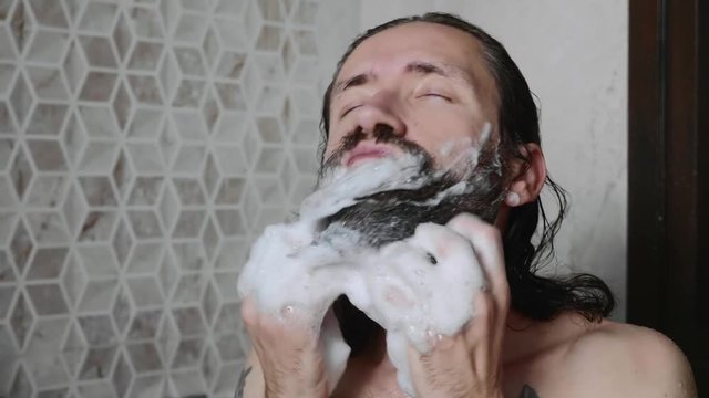 Portrait of young man is washing his beard with shampoo and foam in the shower enjoying the process with closed eyes.