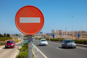 A red white no entry sign with three cars that meet the viewer. The scene has sunshine and blue sky.