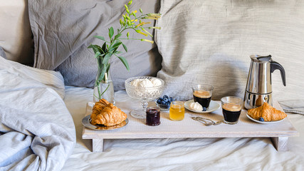 Fototapeta na wymiar Luxury breakfast in the bed in hotel bedroom. Coffee maker and coffee glasses, croissants, jam, raspberry meringue and flowers on wood tray .Good morning surprise concept.