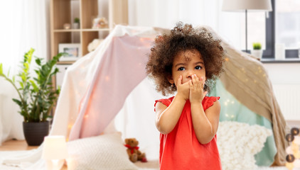 childhood, expressions and emotions concept - confused little african american girl covering mouth...
