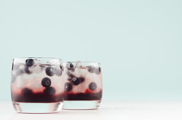 Fresh cold fruit cocktails in two elegant glasses with ice cubes, blueberry on pastel mint color background and white wood table, copy space.
