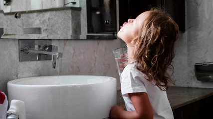 Cute little curly child girl rinses her mouth with water, looking at mirror and spits water into...