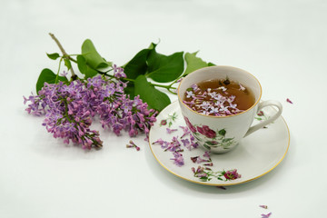 Branch of purple lilac and cup of tea in porcelain. Lilac flowers on the surface of the tea. Copy space. Postcard.