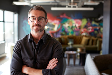 Middle aged white male creative in casual office lounge area looks to camera, arms crossed, waist up