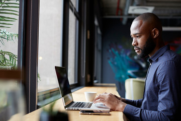 Mid adult black male creative sitting by window in an office social area using a laptop, side view