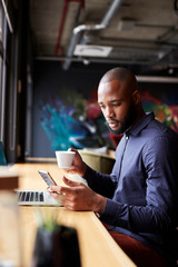 Fototapeta premium Mid adult black male creative sitting by window having coffee using smartphone, side view, vertical