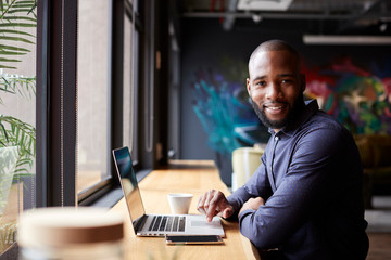 Mid adult black male creative sitting by window in cafe using laptop, turning and smiling to camera