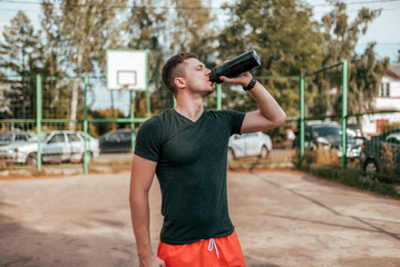 Male athlete, in the summer in the city on the sports ground. In hand a shaker with water and protein. Drinks, resting after a workout fitness in nature. The concept of quenching thirst. Casual wear.