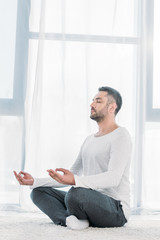 handsome man with eyes closed sitting on carpet in Lotus Pose and meditating at home