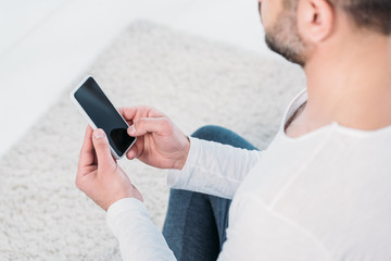 cropped view of man in casual clothes sitting on carpet and using smartphone with blank screen at home