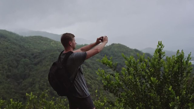 A traveler with a backpack on his back, stands on top of a green hill and takes a panorama of a beautiful landscape on a mobile phone.