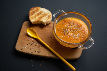 on a black background pumpkin soup in a transparent glass plate, on a wooden board a piece of bread and a yellow spoon, sesame and flax seeds, top view