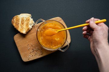 a young girl's hand is holding a yellow spoon submerged in pumpkin soup, a wooden board on a black background, a piece of bread, sesame and flax seeds