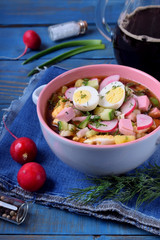 Russian cold soup Okroshka with vegetables, sausage, eggs and kvass in a ceramic bowl on the blue table