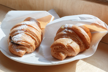 two fresh croissants sprinkled with icing sugar in white paper on a plate. croissants with chocolate are on the windowsill, selective focus.