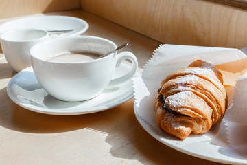 Traditional freshly baked croissant with chocolate and cup of coffe on a wooden window sill in sun rays, selective focus.