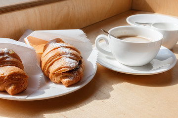 fresh baked croissants and white Coffee cup on wooden background. mug of coffee, croissants with icing sugar wrapped in paper, selective focus