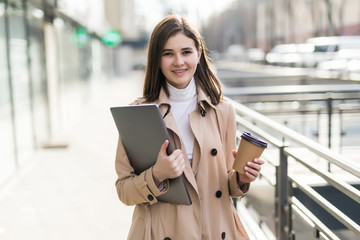 Portrait of joyful european woman walking against office building outdoor with silver laptop and takeaway coffee in hands