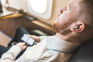 Fototapeta premium Young attractive and successful businessman is resting and listening to music works while sitting in the chair of his private business plane