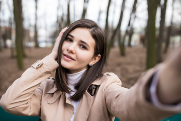 Fototapeta premium Attractive young woman wearing coat sitting on the bench outdoors, taking a selfie